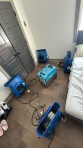 Water damage restoration drying equipment in a bedroom, with multiple air movers and a dehumidifier set up to dry hardwood flooring after a leak.