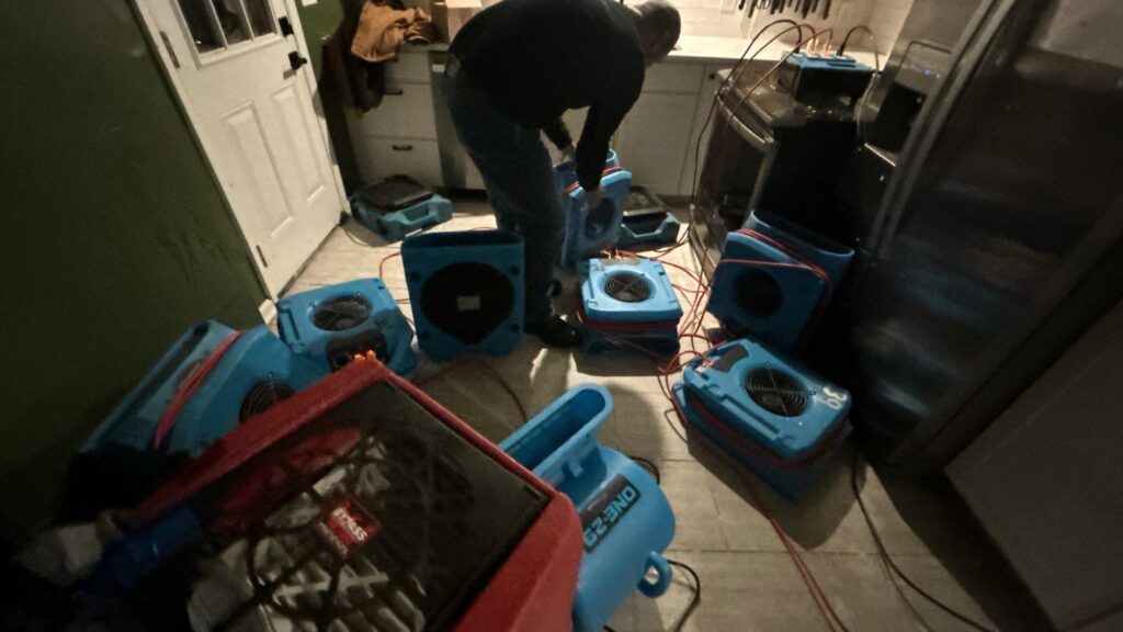 Technician setting up drying equipment after water damage, multiple air movers and dehumidifier running indoors