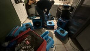 Technician setting up drying equipment after water damage, multiple air movers and dehumidifier running indoors